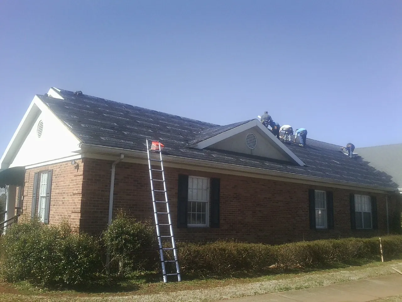 Crew tearing shingles off a brick ranch roof with a ladder set — Emergency Roof Repair work by our crew in Rhine