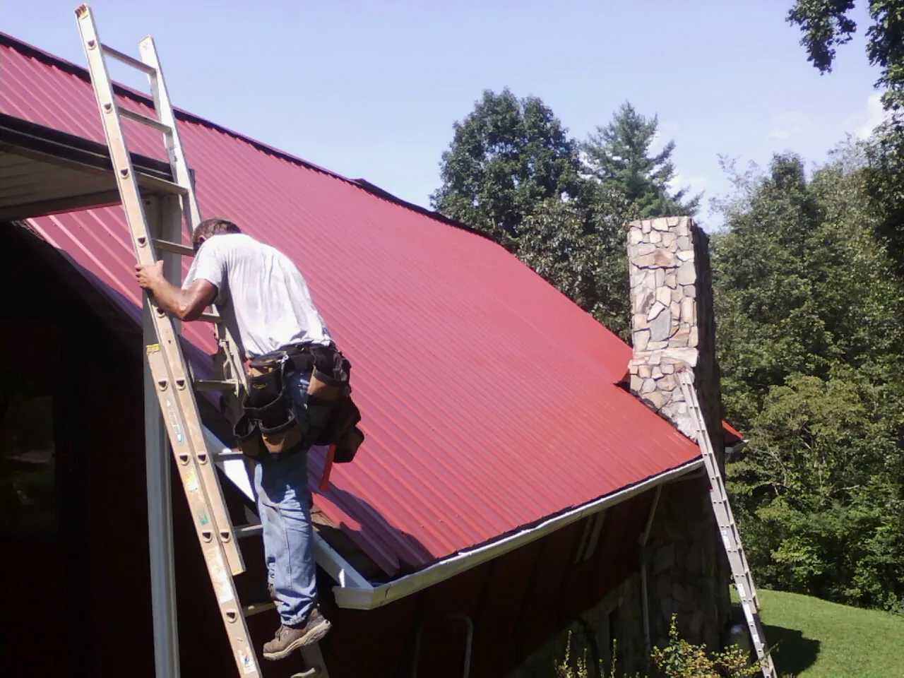 Roofer on a ladder beside a red metal roof and stone chimney — Soffit & Fascia Repair by Roofing Brothers in Rhine